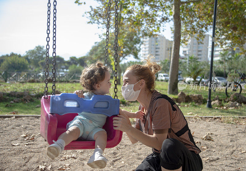 在新的正常生活中，小女孩和妈妈在公园里荡秋千。为了预防冠状病毒感染，母亲戴着防护口罩。