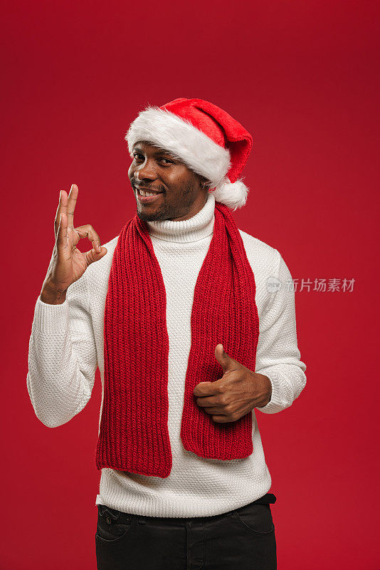 一个皮肤黝黑的男人，穿着羊毛衫，戴着圣诞帽，微笑着表示ok