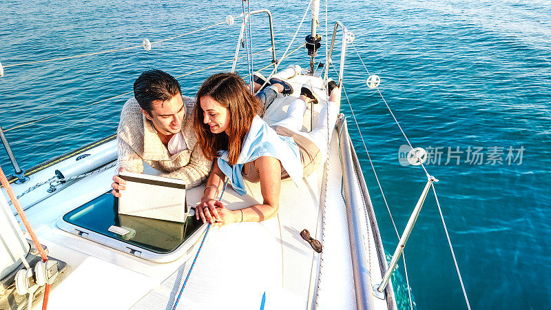 相爱的年轻夫妇在帆船上享受平板电脑的乐趣-豪华旅行生活方式和数字游牧概念的专属游艇旅游-明亮生动的滤镜与软鱼眼镜头失真