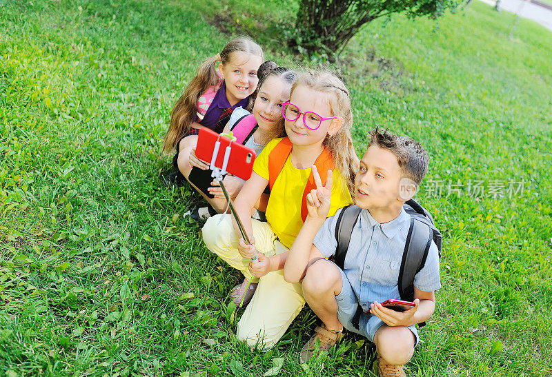 一群学生坐在草地上，穿着五颜六色的衣服，背着书包，微笑着玩耍，用自拍杆上的智能手机相机拍照。回到学校，电子产品，智能手机成瘾