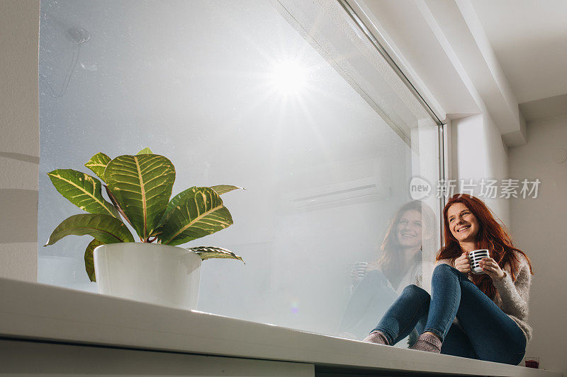 快乐的红发女人在家喝咖啡时在窗边放松。