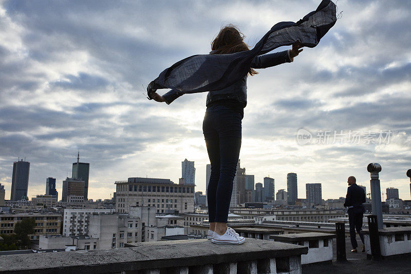 伸开双臂迎着风。在屋顶上保持平衡的漂亮女人。拿着围巾