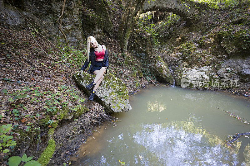 美丽，性感的金发女孩坐在岩石旁边的一个小池塘和古老的石桥