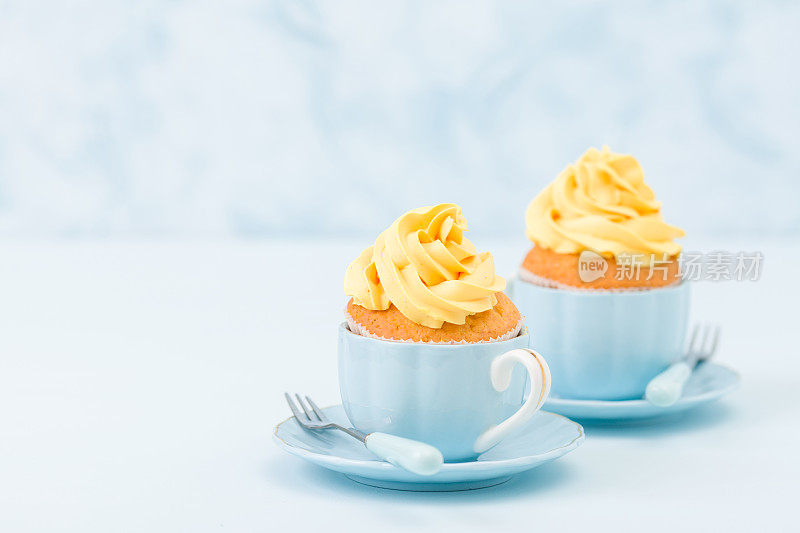 蓝色粉彩背景上的两个小杯子上有甜黄色奶油装饰的纸杯蛋糕。