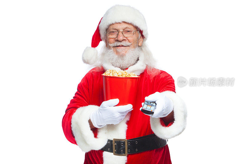 圣诞节。善良的圣诞老人面带微笑，戴着白手套，一只手拿着一桶红色的爆米花，另一只手拿着电视遥控器，切换频道。带着爆米花看电影的概念。孤立在白色背景上。