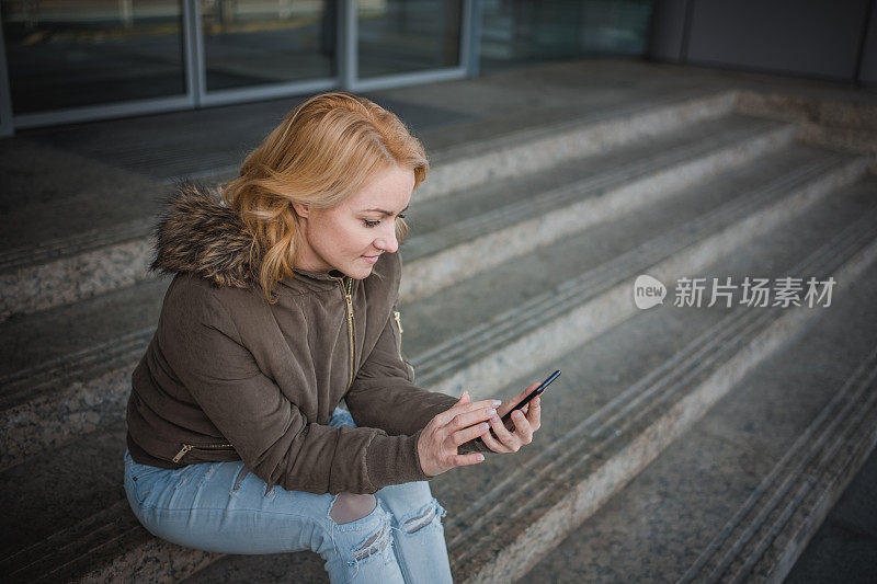 微笑的女人一边在户外的台阶上放松，一边用手机发短信。