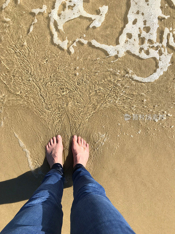 光着脚的男人两只白脚站在海滩上划着海浪，溅起潮水的涟漪，光着脚在潮湿下沉的沙子上留下脚印，海水的水滴和湿牛仔裤溅起水，沙子被冲走，泡沫