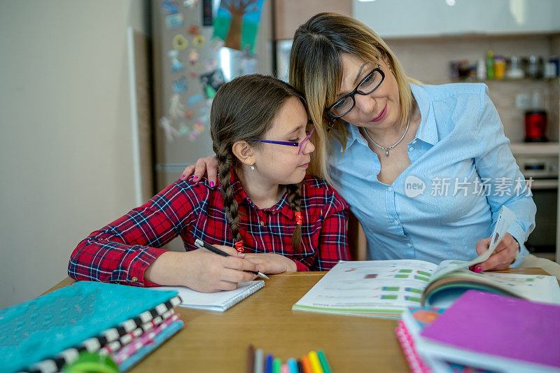 迷人的年轻女子和她可爱的小女儿正坐在桌旁一起做作业。母亲帮助女儿学习学校课程。