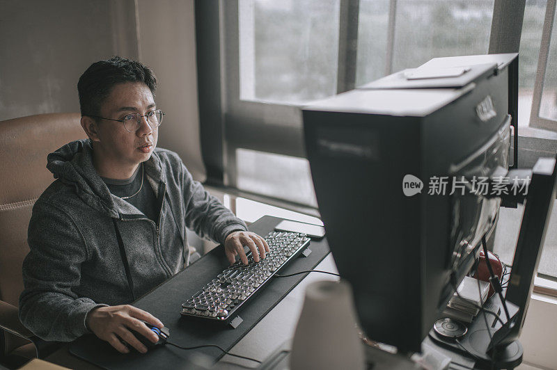 一个亚洲华人中年男子在他的家庭办公室在台式电脑前工作
