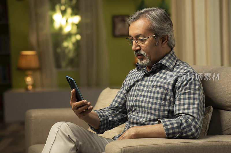 一个成熟的男人坐在家里的沙发上使用手机的照片:库存照片