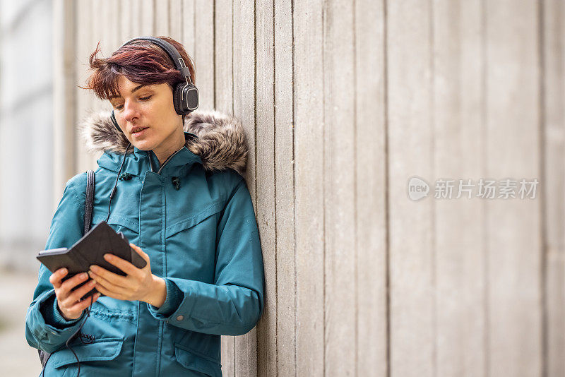 一名身穿蓝色雨衣的年轻女子倚靠在城市街道外的一堵混凝土墙上，查看她的手机