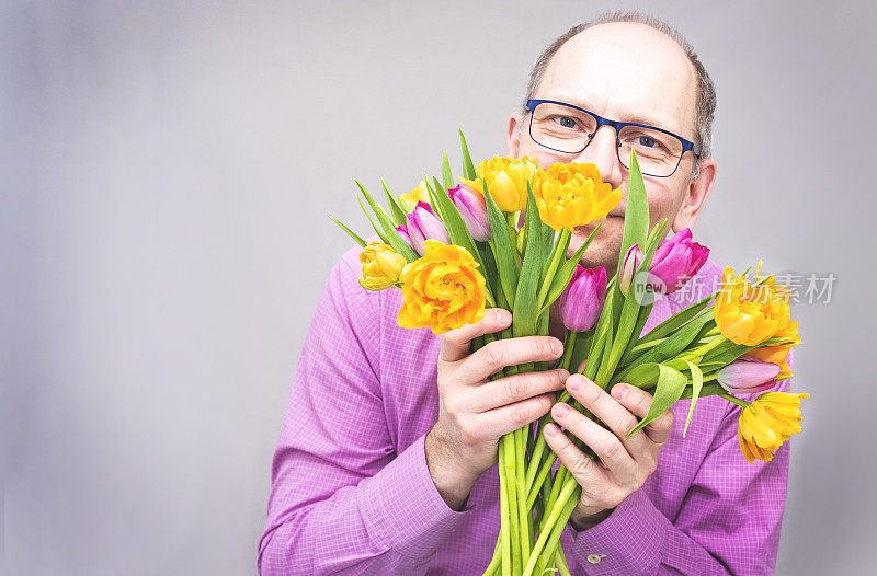 一名白人男子捧着一束灿烂的春天的鲜花，手里拿着一束郁金香，微笑着。一大束黄色和紫色的郁金香。