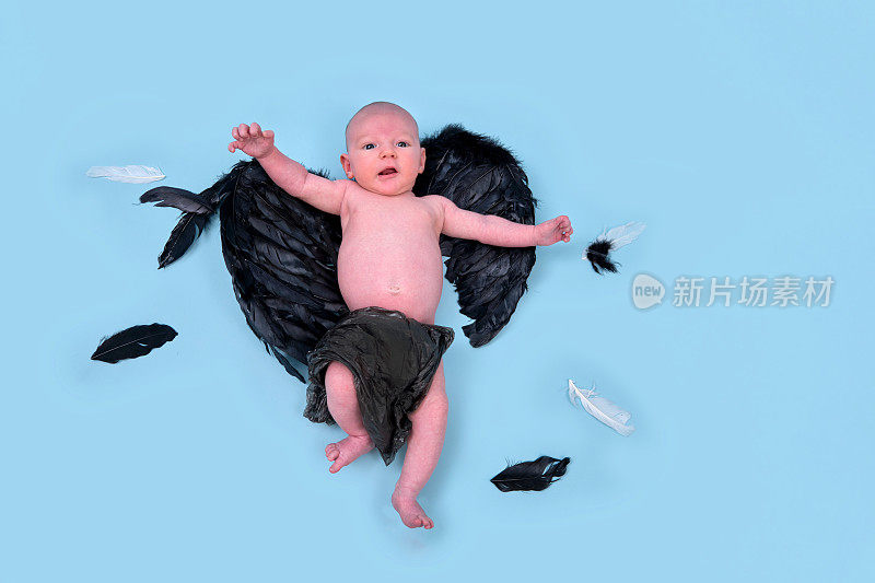 一个长着黑色邪恶翅膀的新生男婴在一个蓝色的工作室背景上，复印空间。一个穿着尿布的白人婴儿一个月大