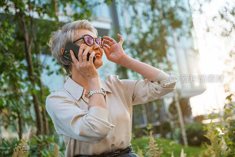 一位身着米色衬衫、面带微笑的女商人正调整着眼镜、在讲电话，她坐在街道上，身后是商业中心和树木，