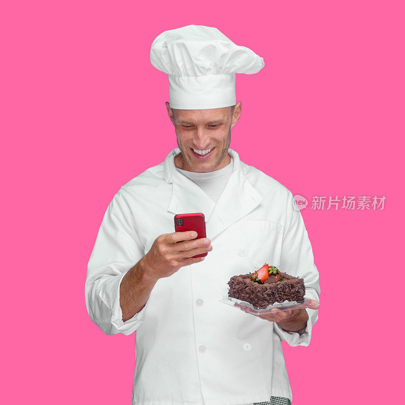 年轻的白人男厨师站在有色背景前，穿着夹克，拿着蛋糕，用手机发短信
