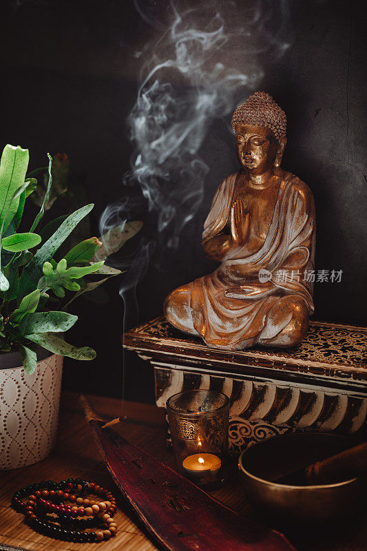 低调的静物画像，一个金佛雕像与冒烟的熏香，点燃的蜡烛，一个唱歌的碗，念珠和一个绿色的植物在豆荚