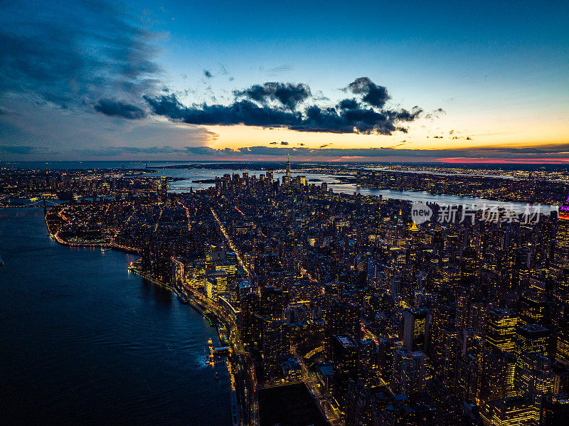 河流环抱的城市夜景航拍，令人屏息。明亮的窗户和五颜六色的夕阳天空。美国纽约曼哈顿