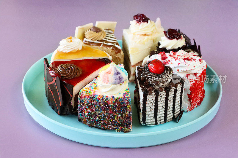 粉彩蓝色盘子上的各种蛋糕切片特写图像，天使食物蛋糕，巧克力和樱桃蛋糕，黑森林蛋糕，红丝绒蛋糕，彩虹蛋糕，咖啡和大理石巧克力蛋糕，咸味焦糖蛋糕，紫色背景，重点在前景