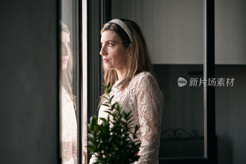 白人成熟女子在白色连衣裙和发带在她的头发站在窗外平静和放松旁边的植物