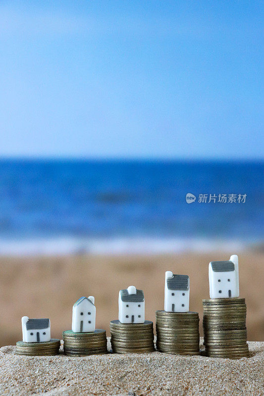 图片:一排度假别墅模型站在成堆的硬币上，沙滩背景与海浪在退潮时破碎，大海和海岸线，度假租赁和度假屋概念，重点在前景