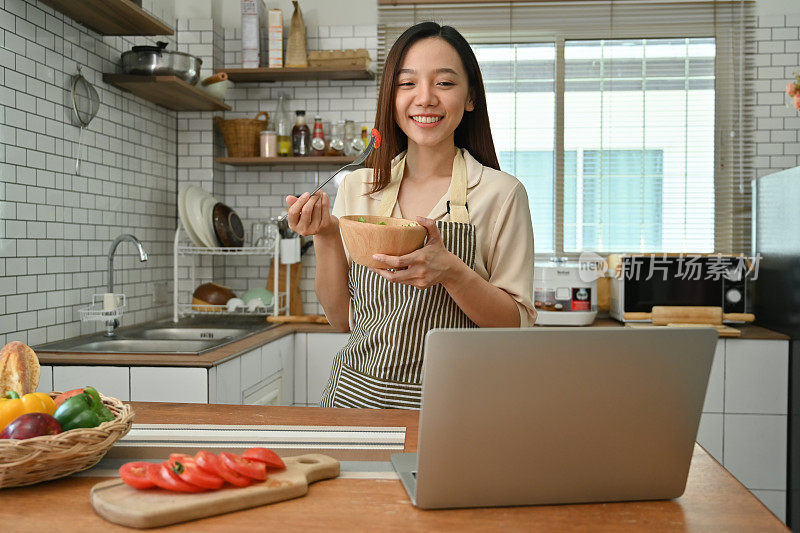 迷人的女人拿着沙拉碗，在家里的厨房里用笔记本电脑进行视频通话。