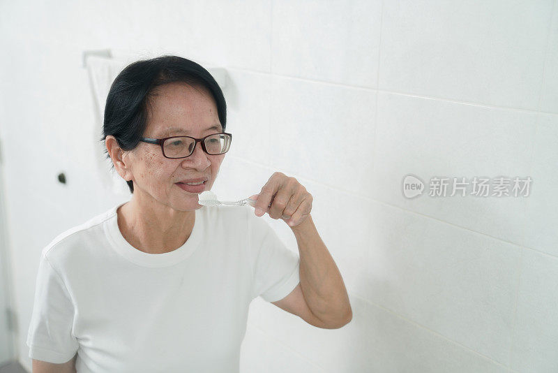 退休的亚洲妇女早上在浴室把牙膏涂在牙刷上。
