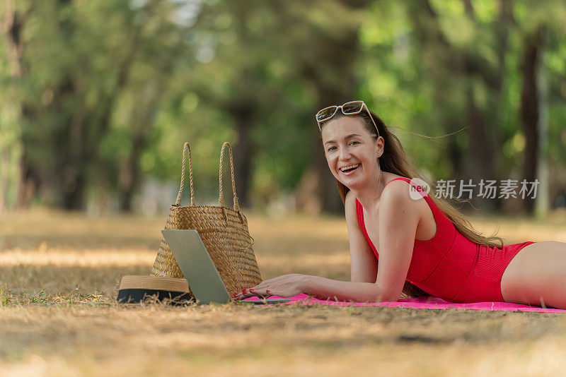 美丽的白人女性性感的女人在红色泳装比基尼戴着太阳镜躺在地上工作在她的电脑笔记本电脑放松在树林里的海滩