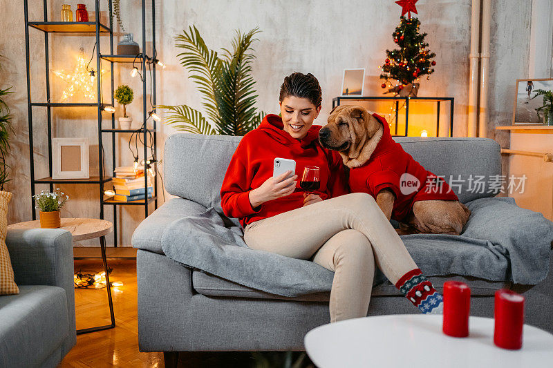 圣诞节时，一位年轻女子和她的狗狗坐在沙发上，一边打电话一边喝酒