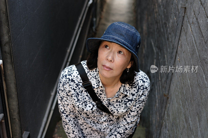 一名戴着帽子的日本妇女站在两栋建筑之间的狭窄小路上，抬起头来