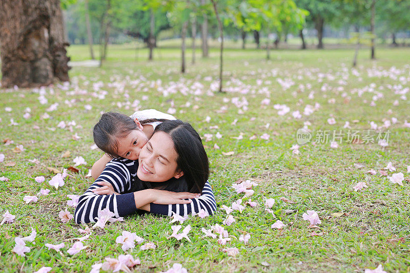 欢快的小女孩依偎着妈妈躺在花园的绿色田野上，粉红色的花已经落满了。幸福美满的家庭。