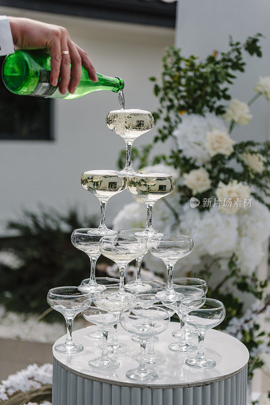 派对上金字塔形的香槟酒杯。男侍、酒保或新郎的手在后院用玻璃杯从瓶子里倒香槟。奢华仪式上的香槟，柔和的焦点。码头庆祝活动