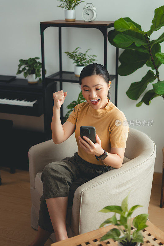 亚洲女性坐在家里的扶手椅上，用智能手机开心兴奋地做出了获胜的手势。