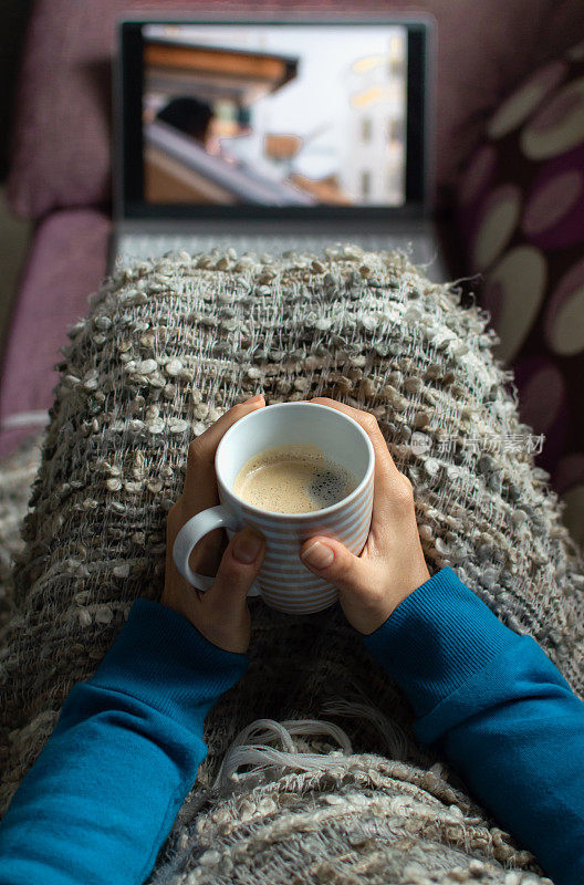 一个女人坐在沙发上，拿着咖啡杯，盖着毯子在笔记本电脑上看电视。