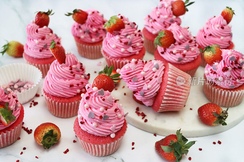 这是一批自制的粉色天鹅绒草莓纸杯蛋糕的图像，草莓上的粉色奶油糖霜呈螺旋状，糖工艺心形和球形点缀，一个纸杯蛋糕躺在一边，从高处看，关注前景