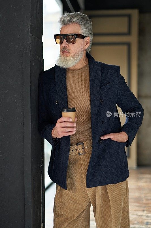 时尚成熟的男人在商务休闲服和太阳镜靠在墙上，拿着一次性咖啡杯，而在阁楼内部摆姿势