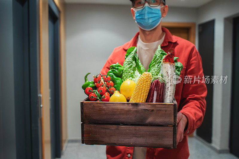 送外卖的人戴着防护口罩，拿着装食物的纸袋站在门口。快递员把装有新鲜蔬菜和水果的盒子交给顾客