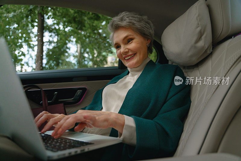 一位微笑的老妇人坐在一辆豪华轿车的副驾驶座位上，正在她的笔记本电脑上打字
