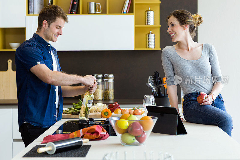 英俊的年轻人打开酒瓶，而他的妻子在厨房使用数字平板电脑。