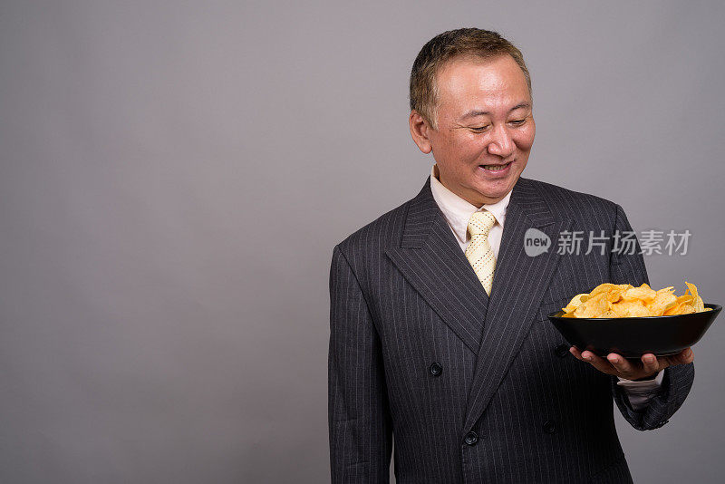 灰色背景下，一名成熟的亚洲商人捧着一碗薯片