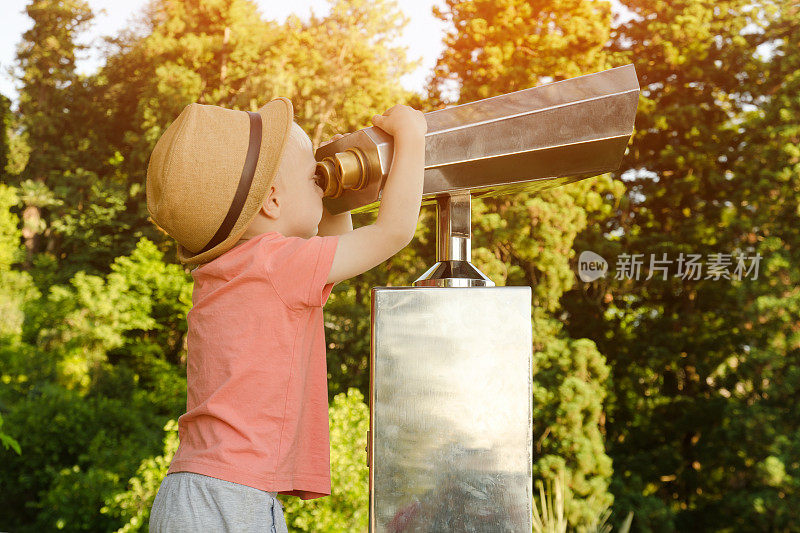 戴着帽子的金发小男孩在看一副大望远镜。背景中的公园