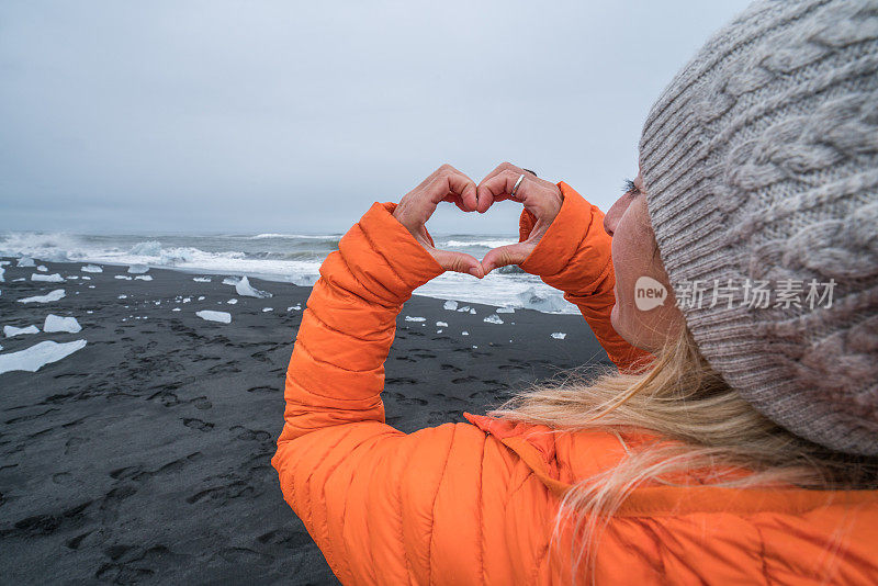 在冰岛有冰山的黑沙滩上，一名年轻女子正在制作心形手指框架