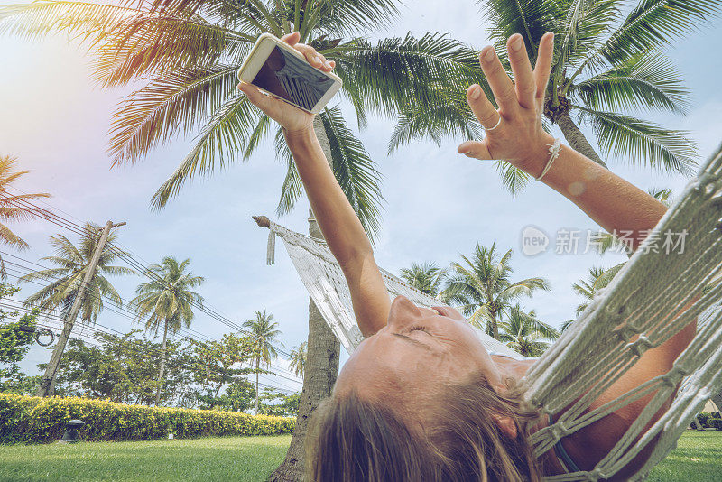 年轻女子在白色吊床上自拍，在热带气候下放松度假——人们在旅行中享受生活和自然，用移动设备技术捕捉凉爽的瞬间