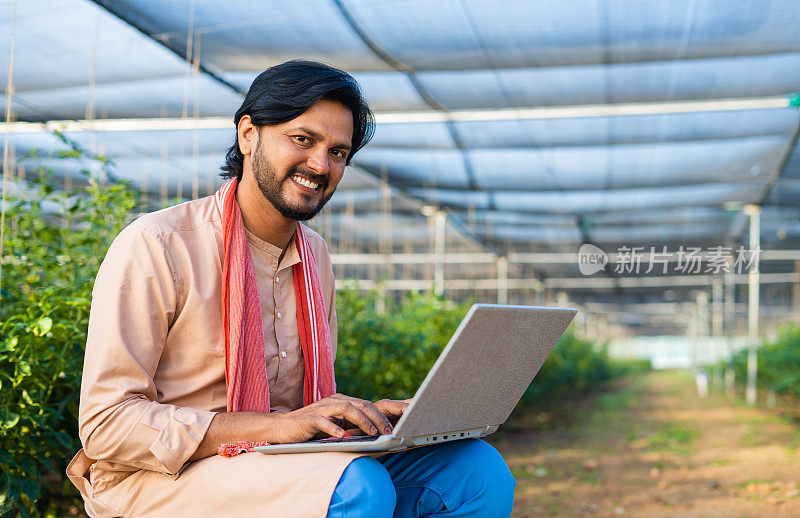 快乐的年轻农民在温室用笔记本电脑工作时看着相机-现代农业，技术和发展或增长的概念。
