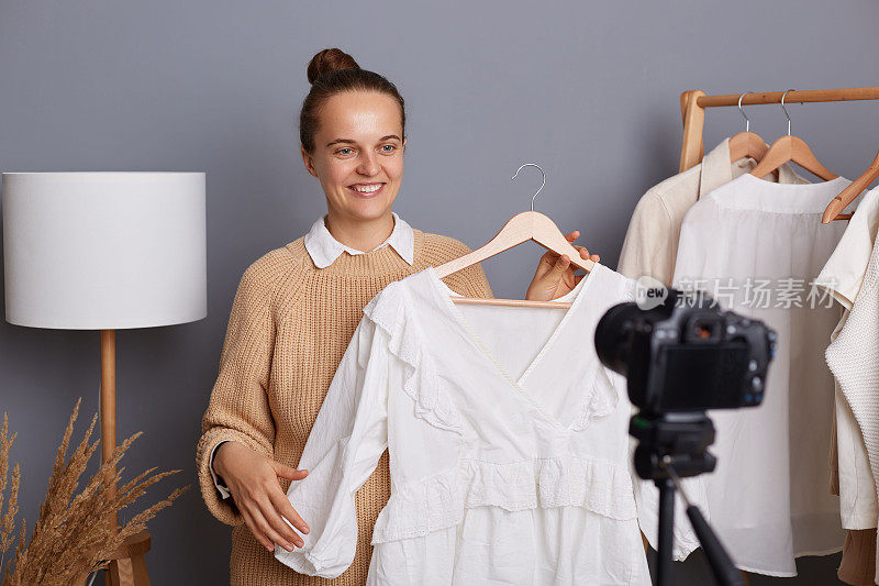 微笑着美丽的白人女子穿着米色毛衣，为vlog拍摄视频，女设计师展示新系列的衬衫，站在灰色的墙上，衣服挂在衣柜的架子上。