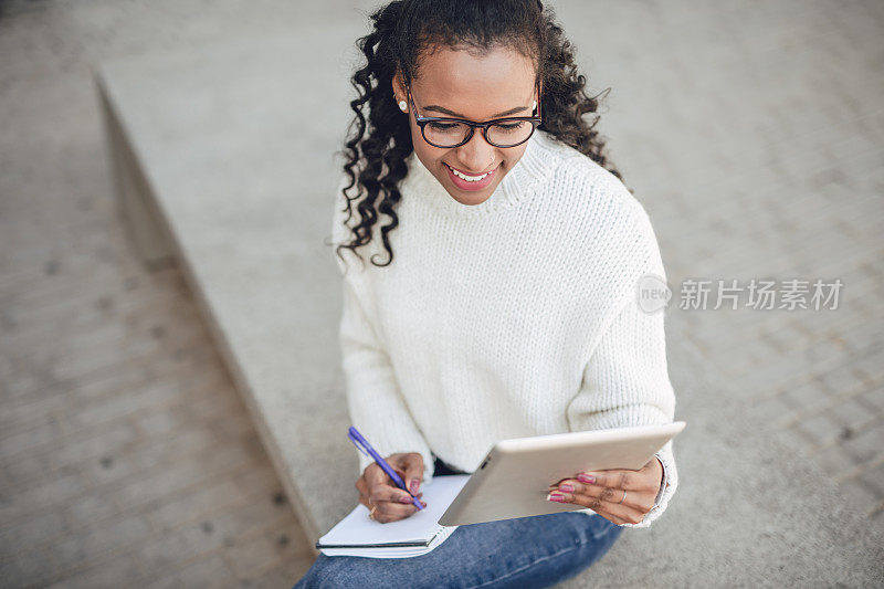 一名身穿白色毛衣的年轻黑人女子坐在公寓大楼前的长凳上，用平板电脑工作