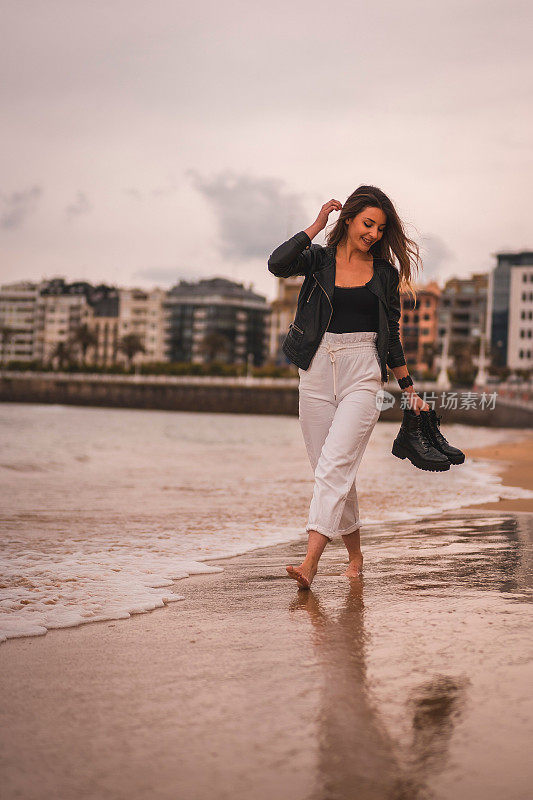 垂直拍摄的年轻时尚的女性在日落时赤脚走在海滩上，抱着她的海岸