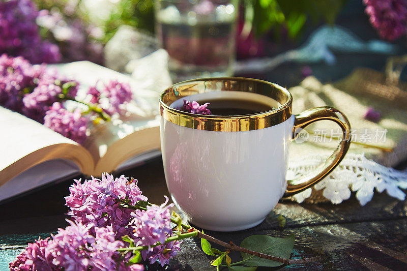 浪漫的阳光花园，一杯咖啡放松休闲。模仿白色杯子和书包围紫丁香花。一杯咖啡，一束丁香花，一本书。硬阴影。夏日，园林唯美