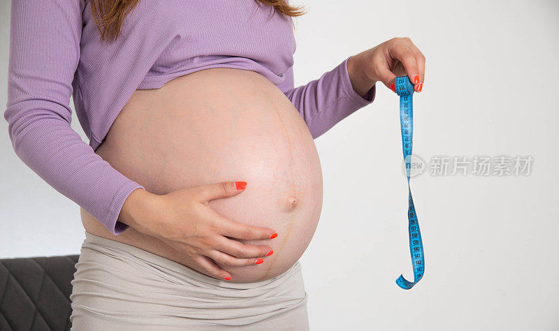 一个大肚子的怀孕女孩拿着白色背景的卷尺。怀孕期间腹部测量和大小的概念。以周为单位的腹部容积。
