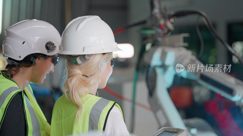 在重工业制造车间，女工程师队伍穿着安全制服，用遥控器控制机械臂焊接工件。技术人员小组检查工厂焊接火点中的自动化机器人系统。