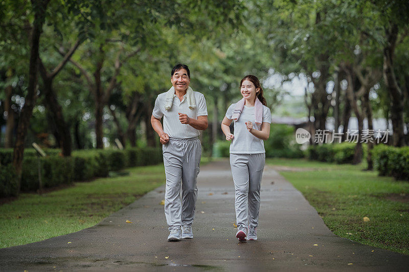 年轻美丽的亚洲女子和她的父亲在公园愉快地慢跑，带着微笑。亚洲女子与父亲慢跑养生，公园运动养生。概念医疗人寿保险。